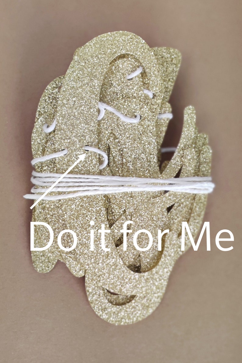 May include: Gold glitter letters spelling "Do it for Me" strung together with white string. The letters are layered on top of each other.