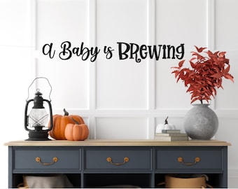 May include: A black wall decal with the text A Baby is BREWING in a decorative font. The decal is displayed above a dark blue console table featuring a black lantern, two orange pumpkins, a white pumpkin, books, and a vase with red leaves.