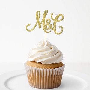 May include: Gold glitter cupcake topper with the letters "M&C" in a cursive font. The topper is on top of a white frosted cupcake.