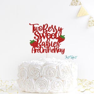 May include: A red glitter cake topper featuring the text Two Berry Sweet Babies Are On The Way with two small strawberry illustrations, positioned on top of a white frosted cake with a rose swirl pattern and gold decorative accents.