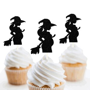 May include: Three black glittery silhouette cupcake toppers featuring a pregnant witch figure holding a broomstick. Each topper is mounted on a thin white stick and is inserted into a cupcake topped with white frosting against a white background.