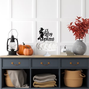 May include: A white cake with a black glitter topper that says "A Baby is Brewing" with a silhouette of a witch on a broomstick. The cake is on a white cake stand and there are pumpkins and a lantern on a dark blue dresser.