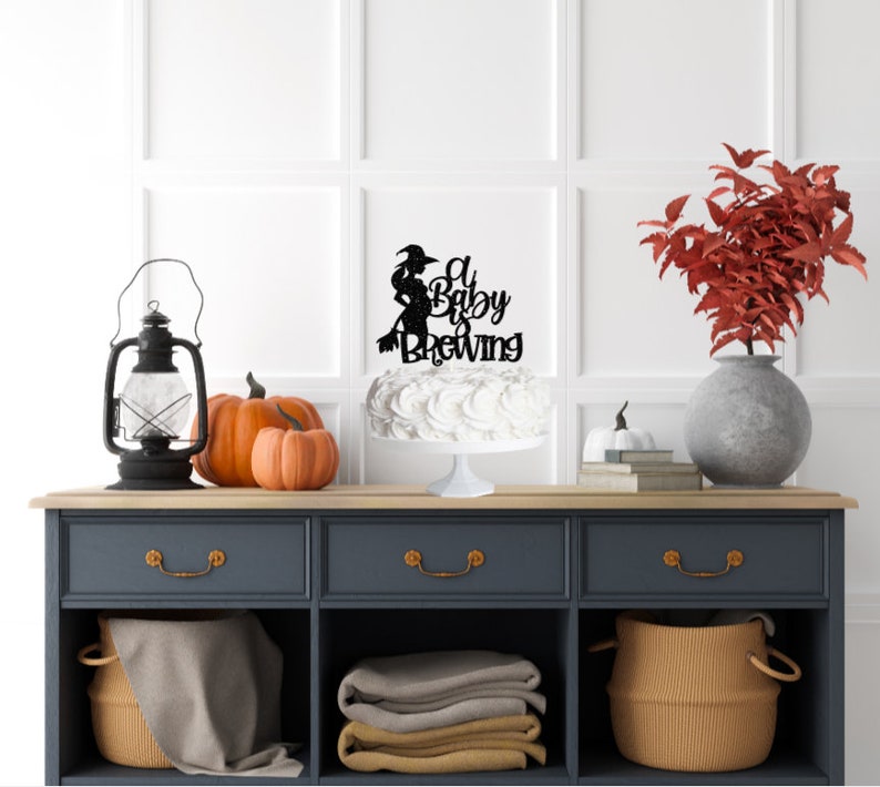May include: A white cake with a black glitter topper that says "A Baby is Brewing" with a silhouette of a witch on a broomstick. The cake is on a white cake stand and there are pumpkins and a lantern on a dark blue dresser.