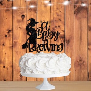May include: A black glitter cake topper with a silhouette of a witch holding a broom and the text "A Baby Is Brewing".