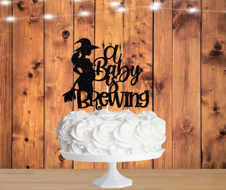 May include: A black glitter cake topper with a silhouette of a witch holding a broom and the text "A Baby Is Brewing".