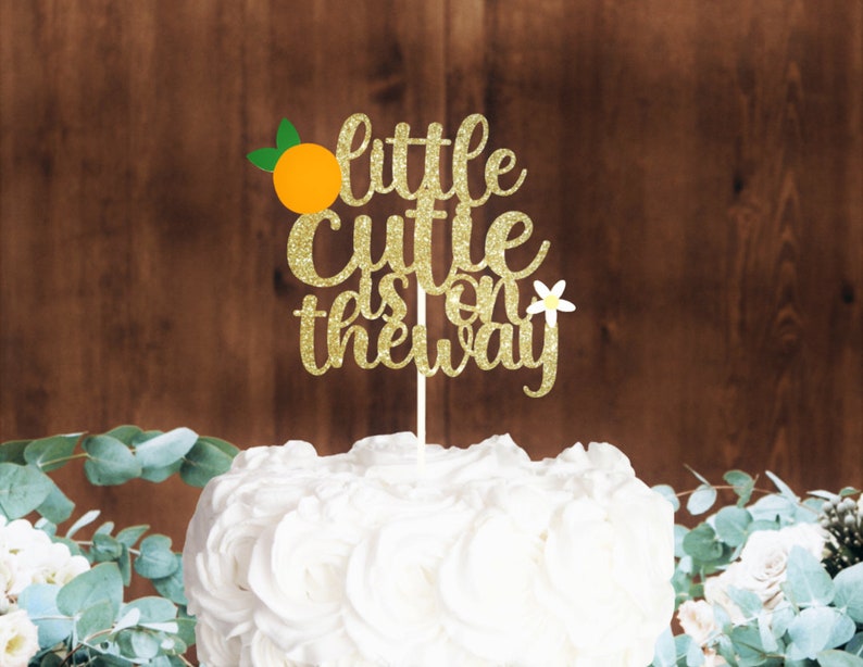 Puede incluir: Topper de pastel de purpurina dorada con el texto "little cutie is on the way" y una rodaja de naranja con hojas verdes. Una flor blanca est&aacute; en el lado derecho del topper.