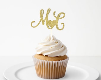 May include: A gold glitter cupcake topper featuring the letters M and L connected by a central heart shape, mounted on a thin stick and inserted into a vanilla cupcake with white frosting in a pleated paper liner on a white plate.