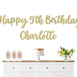 May include: Gold glitter banner with the text "Happy 5th Birthday Charlotte". The banner is hanging above a white dresser with three glass jars filled with candy and a white cake on a cake stand.