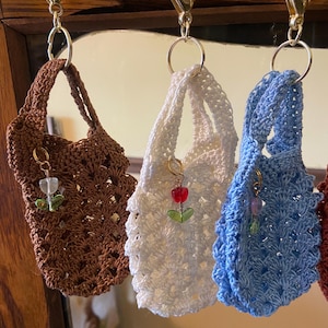 May include: Three miniature crochet tote bag keychains in brown, white, and light blue. Each bag features a textured shell stitch pattern, a single handle, and a small gold-tone lobster clasp. A tiny tulip-shaped bead charm hangs from each bag.