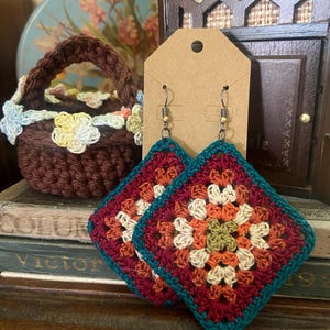 May include: A pair of square crochet earrings with a colorful floral pattern in shades of red, orange, green, and cream, outlined in teal. A small brown crochet basket with floral accents is also visible.