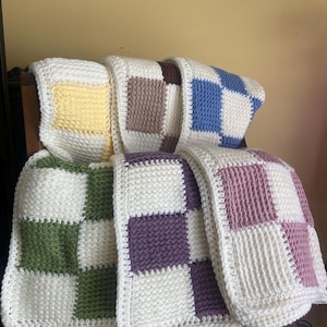 May include: A collection of handmade crochet blankets in various colors, including yellow, brown, blue, green, purple, and pink. Each blanket features a checkerboard pattern with white squares. The blankets are folded and stacked on top of each other.