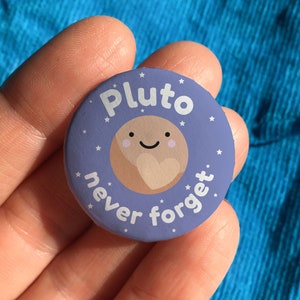May include: A purple button with a cartoon image of Pluto with a heart on it. The text on the button reads "Pluto never forget".