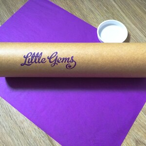 May include: A brown cardboard tube with the text "Little Gems" printed in purple on the side. The tube is sitting on a purple surface.