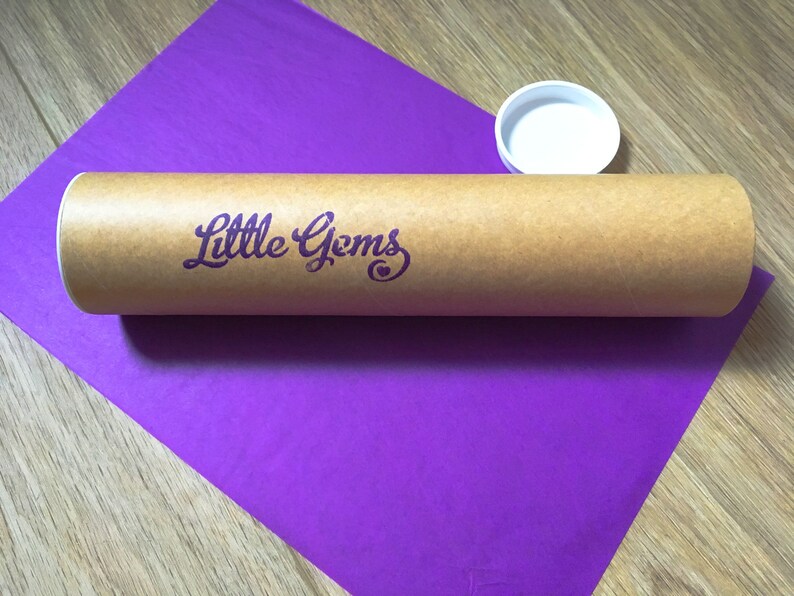 May include: A brown cardboard tube with the text "Little Gems" printed in purple on the side. The tube is sitting on a purple surface.
