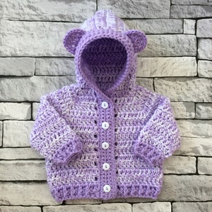 May include: A purple and white striped crocheted baby cardigan with a hood and bear ears. The cardigan has white buttons.