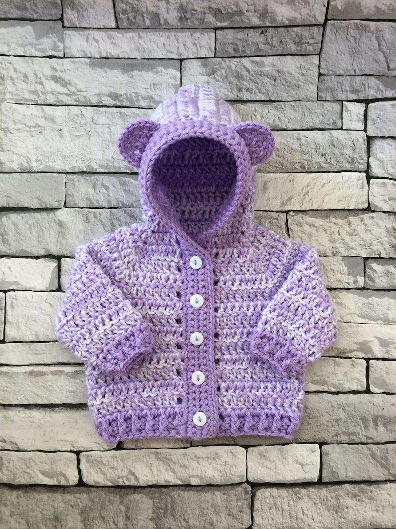 May include: A purple and white striped crocheted baby cardigan with a hood and bear ears. The cardigan has white buttons.