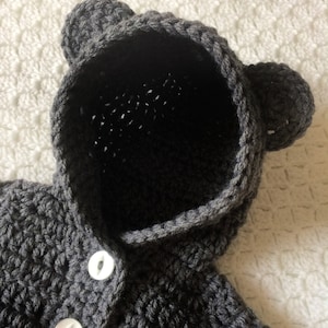 Puede incluir: Un jersey de punto gris con capucha y orejas de oso y botones blancos.