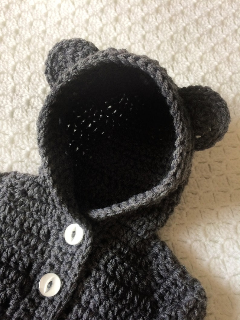 Puede incluir: Un jersey de punto gris con capucha y orejas de oso y botones blancos.