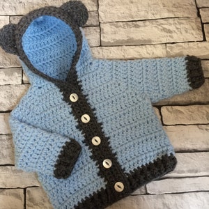 May include: A light blue crocheted baby jacket with a hood and bear ears. The jacket has a gray trim and buttons down the front.