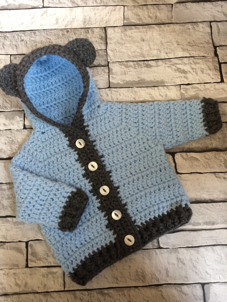 May include: A light blue crocheted baby jacket with a hood and bear ears. The jacket has a gray trim and buttons down the front.