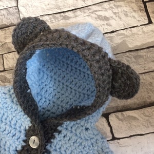 Puede incluir: Un jersey de beb&eacute; de punto de ganchillo azul y gris con capucha y orejeras. El jersey tiene un cierre de bot&oacute;n.
