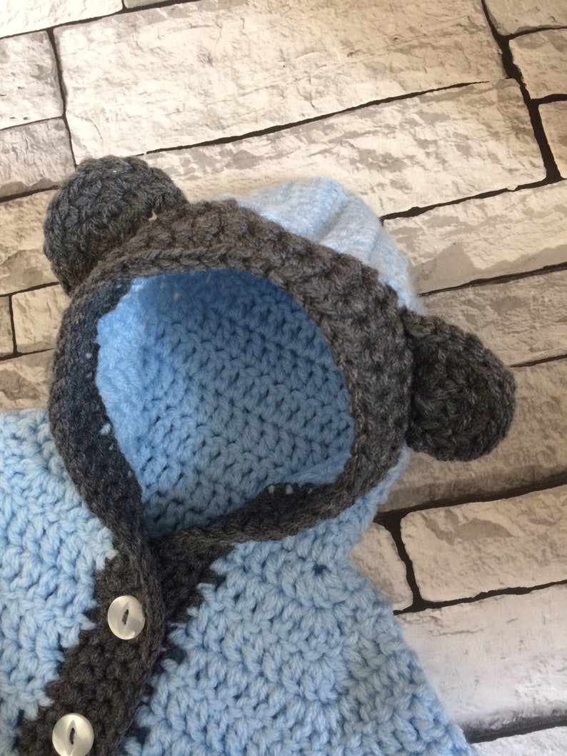 Puede incluir: Un jersey de beb&eacute; de punto de ganchillo azul y gris con capucha y orejeras. El jersey tiene un cierre de bot&oacute;n.