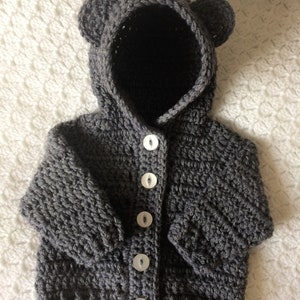May include: A gray crocheted baby cardigan with a bear hood and white buttons.