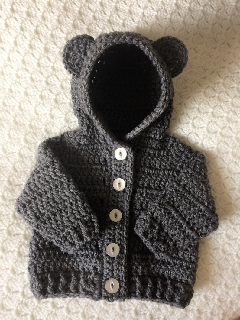 May include: A gray crocheted baby cardigan with a bear hood and white buttons.