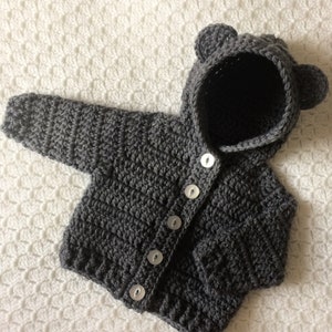 May include: Gray crocheted baby cardigan with a hood and bear ears. The cardigan has a button closure and is made with a soft yarn.