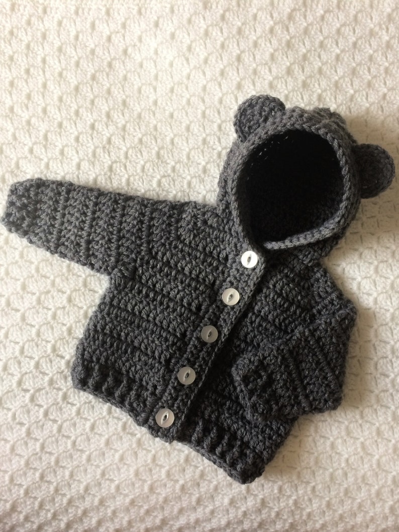 May include: Gray crocheted baby cardigan with a hood and bear ears. The cardigan has a button closure and is made with a soft yarn.