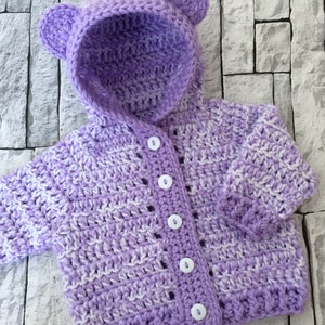 May include: A purple and white striped crocheted baby cardigan with a hood and bear ears. The cardigan has white buttons.