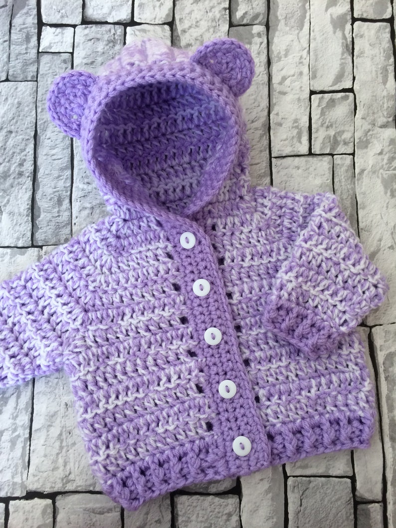 May include: A purple and white striped crocheted baby cardigan with a hood and bear ears. The cardigan has white buttons.