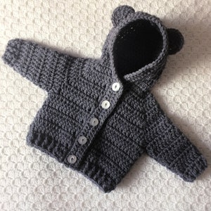May include: Gray crocheted baby cardigan with a hood and bear ears. The cardigan has five white buttons down the front.