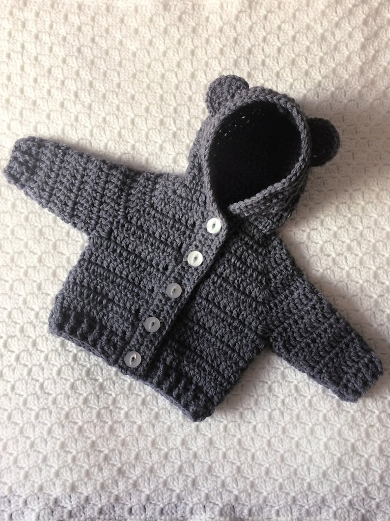 May include: Gray crocheted baby cardigan with a hood and bear ears. The cardigan has five white buttons down the front.