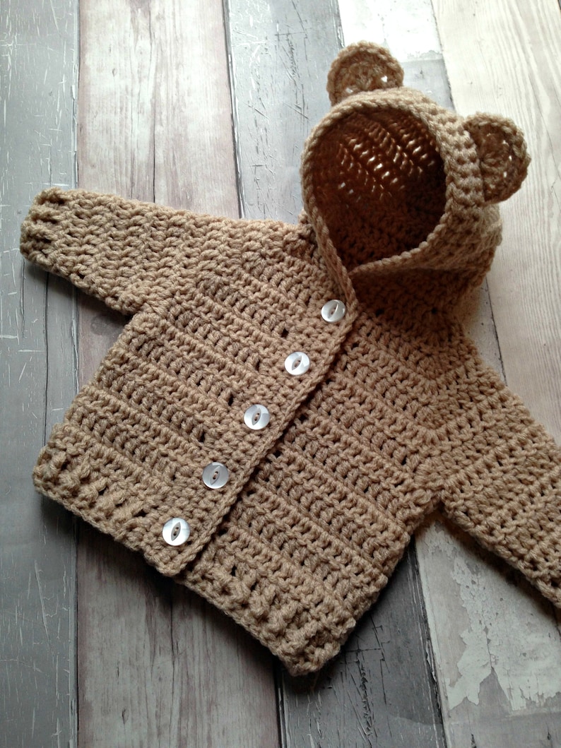 Puede incluir: Un c&aacute;rdigan de beb&eacute; de punto marr&oacute;n con capucha de oso y botones blancos.