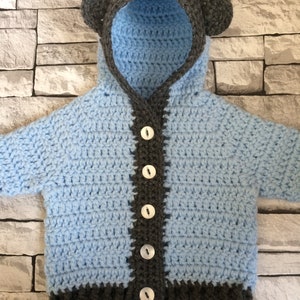 May include: A light blue crocheted baby cardigan with a hood and bear ears. The cardigan has a gray trim and buttons down the front.
