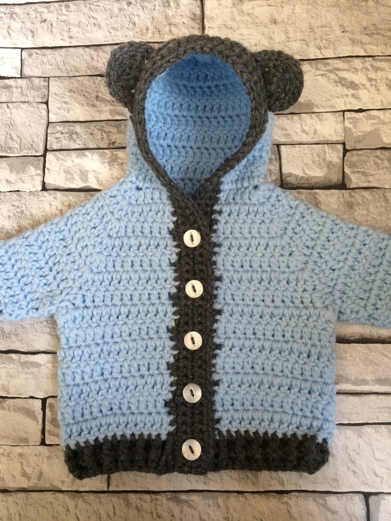 May include: A light blue crocheted baby cardigan with a hood and bear ears. The cardigan has a gray trim and buttons down the front.