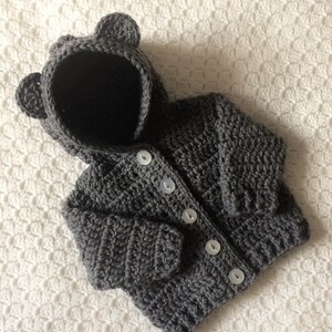 May include: A gray crocheted hooded cardigan with bear ears. The cardigan has a button closure and is made with a soft yarn.