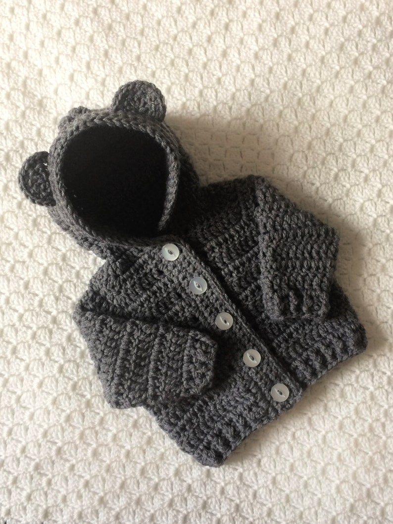 May include: A gray crocheted hooded cardigan with bear ears. The cardigan has a button closure and is made with a soft yarn.
