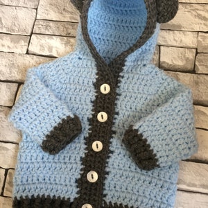 May include: A light blue crocheted baby cardigan with a hood and bear ears. The cardigan has a gray trim and buttons down the front.