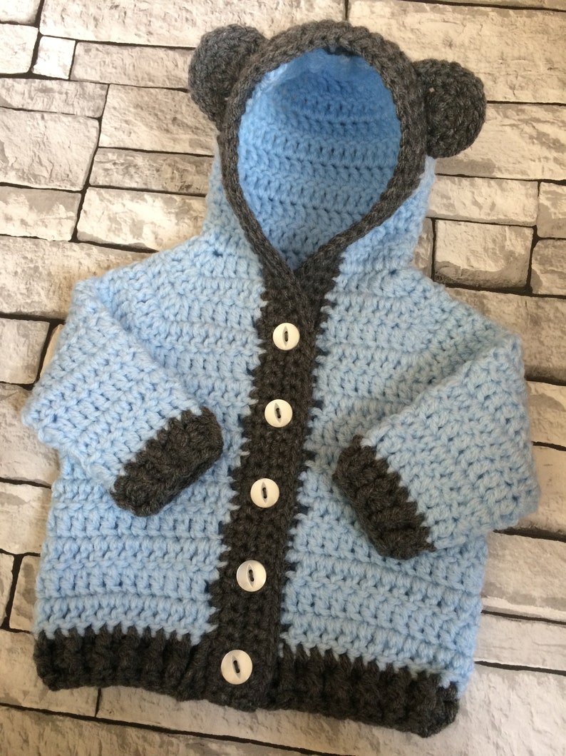 May include: A light blue crocheted baby cardigan with a hood and bear ears. The cardigan has a gray trim and buttons down the front.