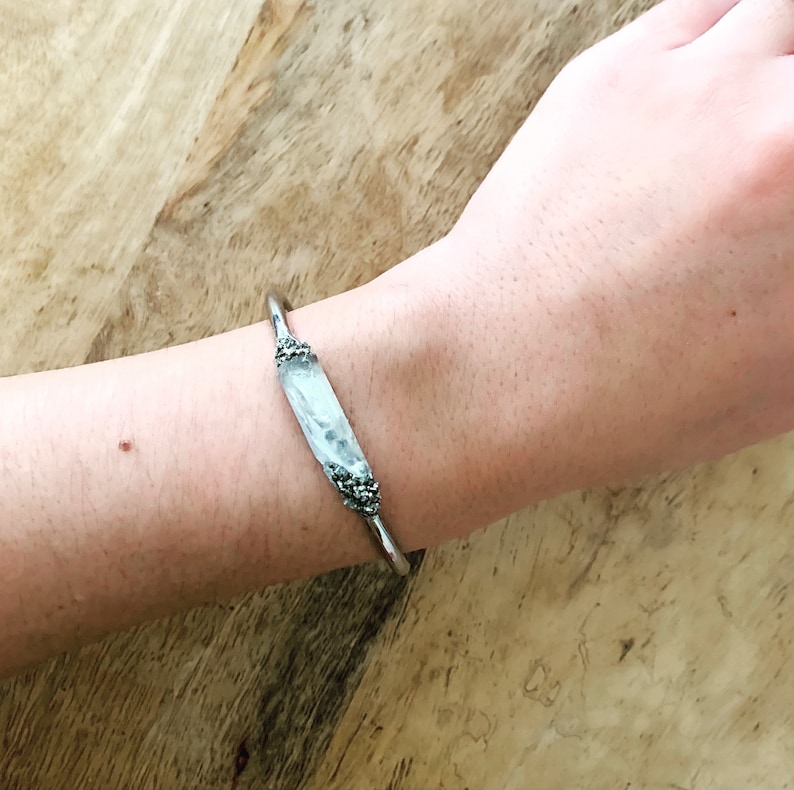 May include: A silver bangle bracelet with a white, textured, rectangular stone set in the center. The stone is surrounded by small silver beads.