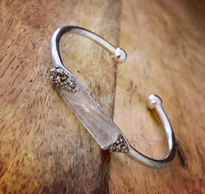 May include: A silver bangle bracelet with a clear crystal bar set in the center. The crystal bar is surrounded by small silver beads.