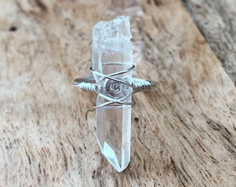 May include: A clear quartz crystal wrapped in silver wire, forming a ring. The crystal is pointed at the top and has a natural, rough texture.