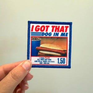 May include: A square embroidered patch with a blue border featuring the red and blue text I GOT THAT DOG IN ME above an image of a hot dog and a blue drink cup. The bottom section includes the text 1/4 POUND PLUS ALL BEEF HOT DOG AND 20 oz. SODA.