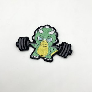 May include: Embroidered patch featuring a cartoon triceratops lifting a barbell. The dinosaur is green, yellow, and white with black accents. The barbell is black. The patch is likely for clothing or accessories.