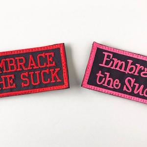May include: Two rectangular embroidered patches. One features the phrase "Embrace the Suck" in red on a black background with a red border. The other has pink text and border.