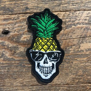 May include: A black and white embroidered skull wearing sunglasses with a yellow pineapple on top. The pineapple has green leaves.