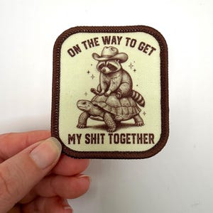 May include: A rectangular patch with a brown border and a light green background. The patch features a raccoon wearing a cowboy hat riding a turtle. The text "ON THE WAY TO GET MY SHIT TOGETHER" is printed in brown.