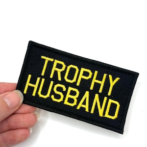 May include: A black rectangular patch with yellow embroidered text that reads "TROPHY HUSBAND". The patch is held against a white background. The text is in a bold, sans-serif font.